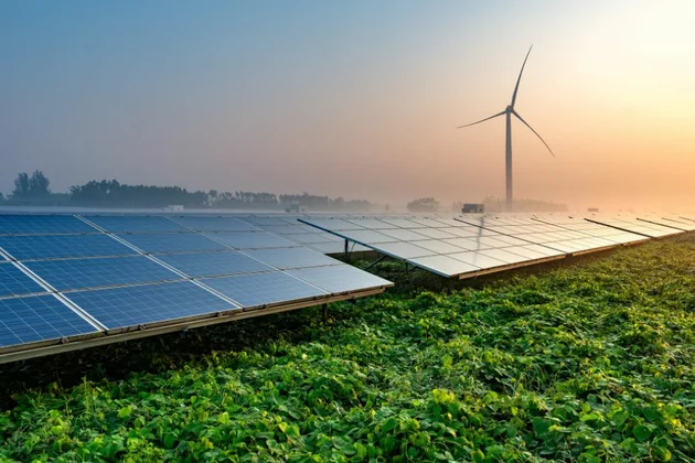 auf einer Rasenfläche befinden sich Fotovoltaikanlagen hinter ihnen vor einer untergehenden Sonne ein Windrad